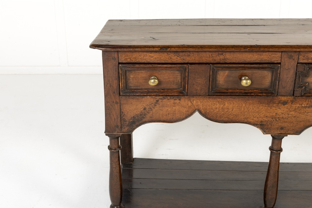 An 18th-century dark oak dresser base featuring geometric panelled drawer fronts, a shaped lower apron, turned front legs, and a bottom potboard shelf. View VIII