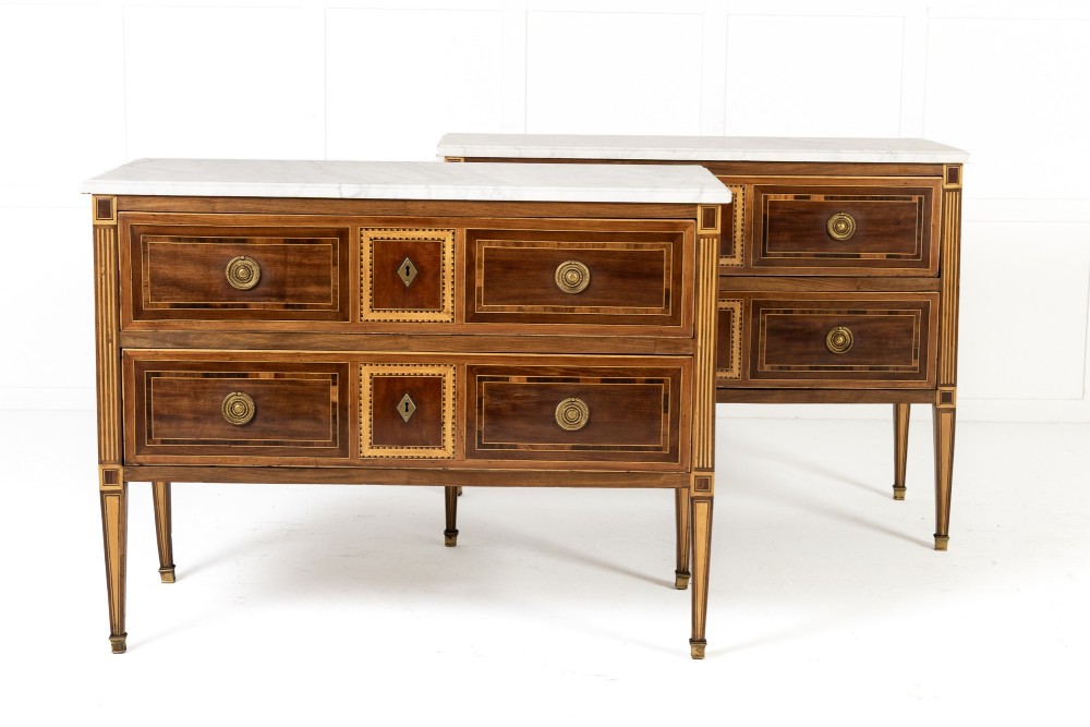A pair of 18th-century Italian Neoclassical chests of drawers featuring geometric wood inlays, brass ring handles, light marble tops, and tapered square legs.