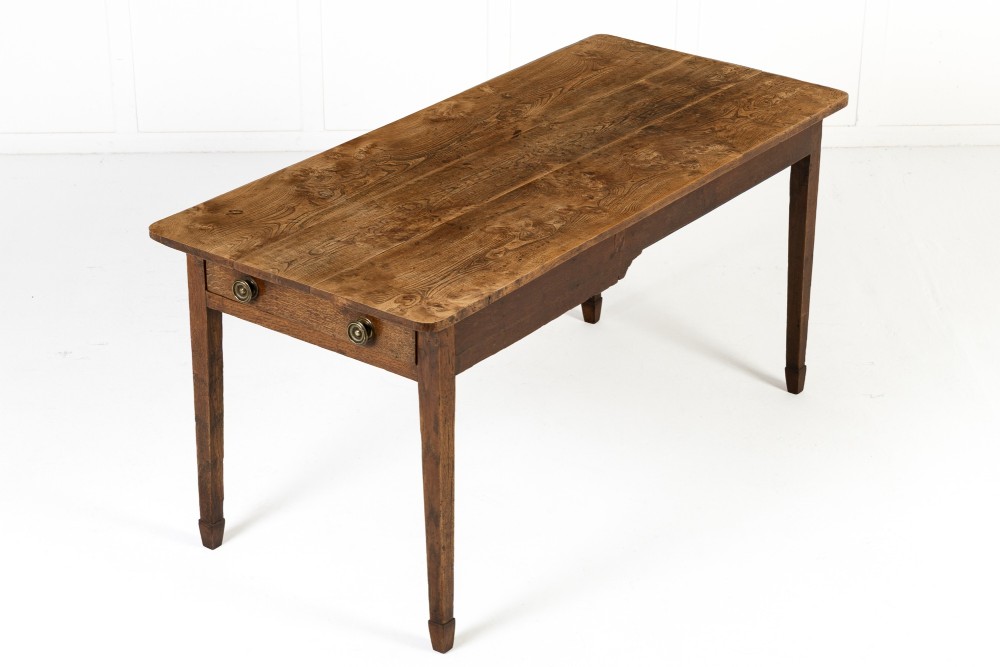 An 18th-century English elm farmhouse table featuring a rectangular plank top with prominent wood grain, a side drawer with brass knobs, and tapered square legs.