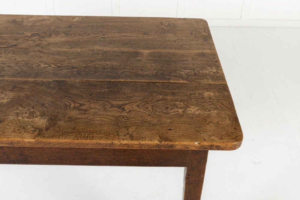 An 18th-century English elm farmhouse table featuring a rectangular plank top with prominent wood grain, a side drawer with brass knobs, and tapered square legs. View IV