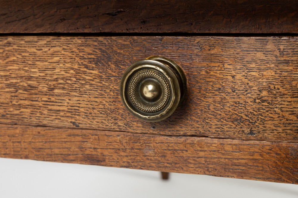 An 18th-century English elm farmhouse table featuring a rectangular plank top with prominent wood grain, a side drawer with brass knobs, and tapered square legs. View V