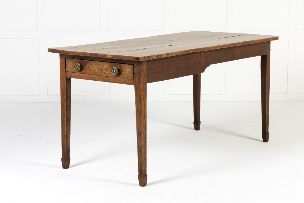 An 18th-century English elm farmhouse table featuring a rectangular plank top with prominent wood grain, a side drawer with brass knobs, and tapered square legs. View VI