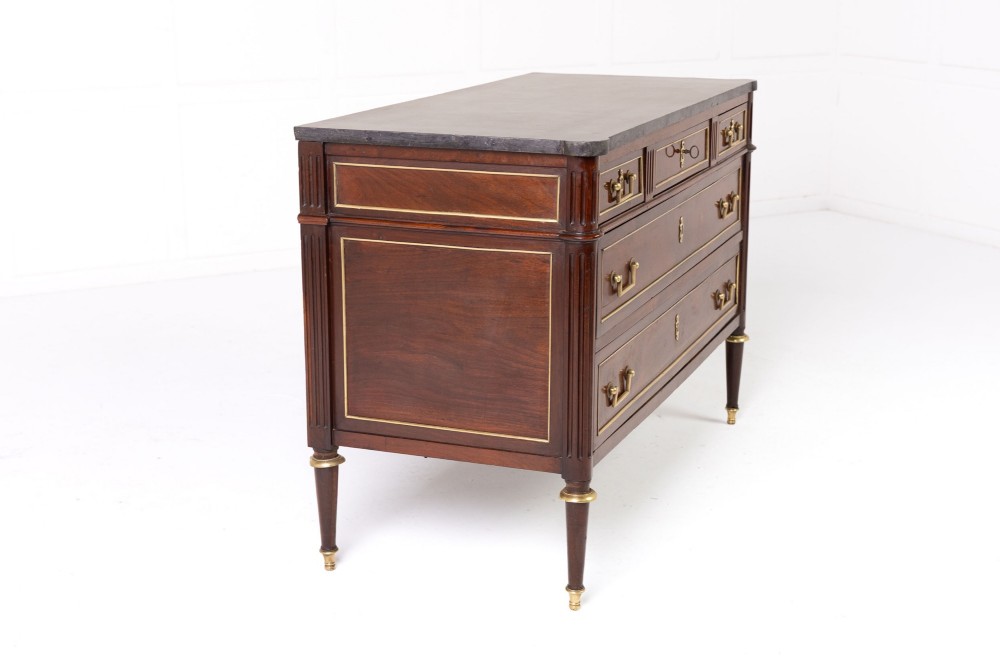 An 18th-century French mahogany commode featuring three upper short drawers, two long lower drawers with brass moulding, turned fluted legs, and a dark marble top. View IV