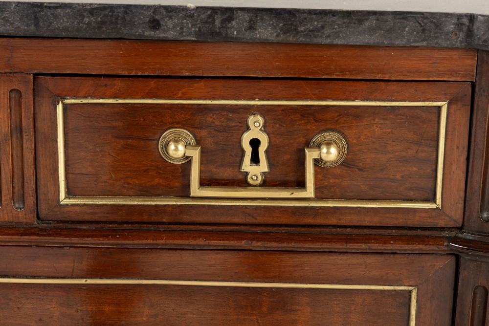An 18th-century French mahogany commode featuring three upper short drawers, two long lower drawers with brass moulding, turned fluted legs, and a dark marble top. View VII