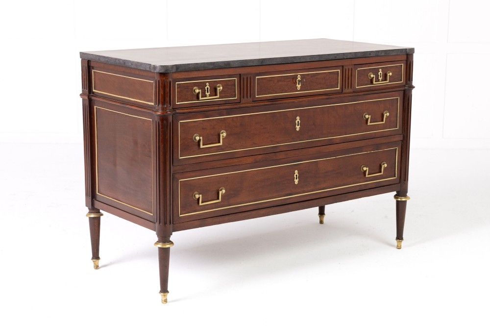 An 18th-century French mahogany commode featuring three upper short drawers, two long lower drawers with brass moulding, turned fluted legs, and a dark marble top. View IX