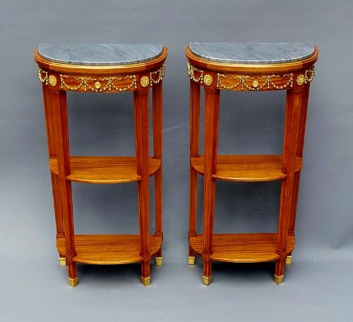 fig 1. A pair of French Louis XVI style demilune side tables featuring grey marble tops, open two-tiered wooden shelving, and ornate gilt bronze floral garland mounts.