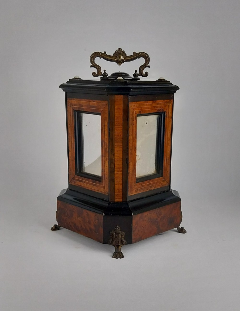 An upright Victorian hexagonal musical cigar box featuring ebonised wood, burr walnut panels, glass inserts, an ornate brass top handle, and brass claw feet.