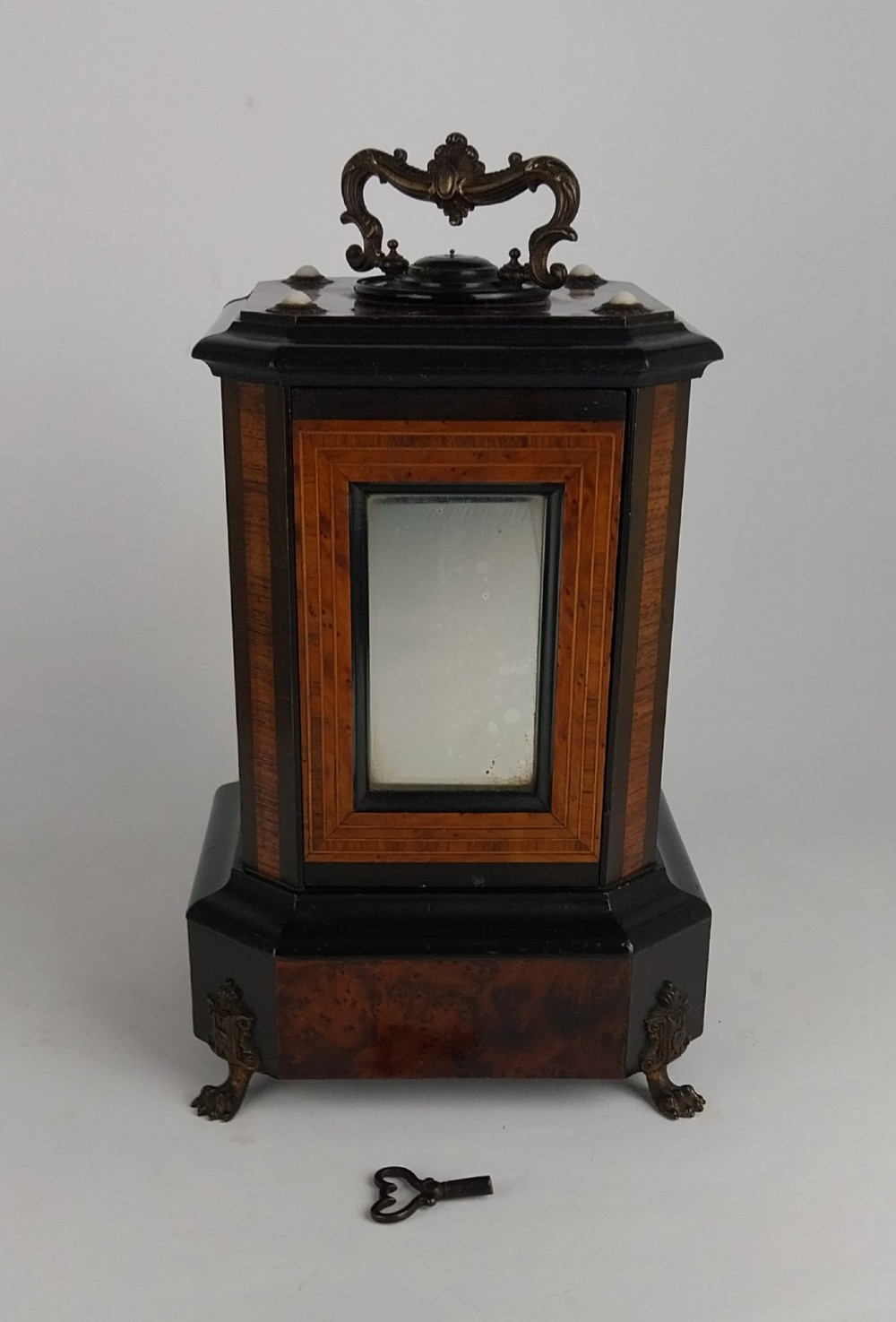 An upright Victorian hexagonal musical cigar box featuring ebonised wood, burr walnut panels, glass inserts, an ornate brass top handle, and brass claw feet. View IV