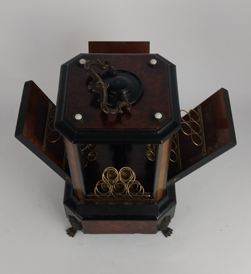 An upright Victorian hexagonal musical cigar box featuring ebonised wood, burr walnut panels, glass inserts, an ornate brass top handle, and brass claw feet. View VI