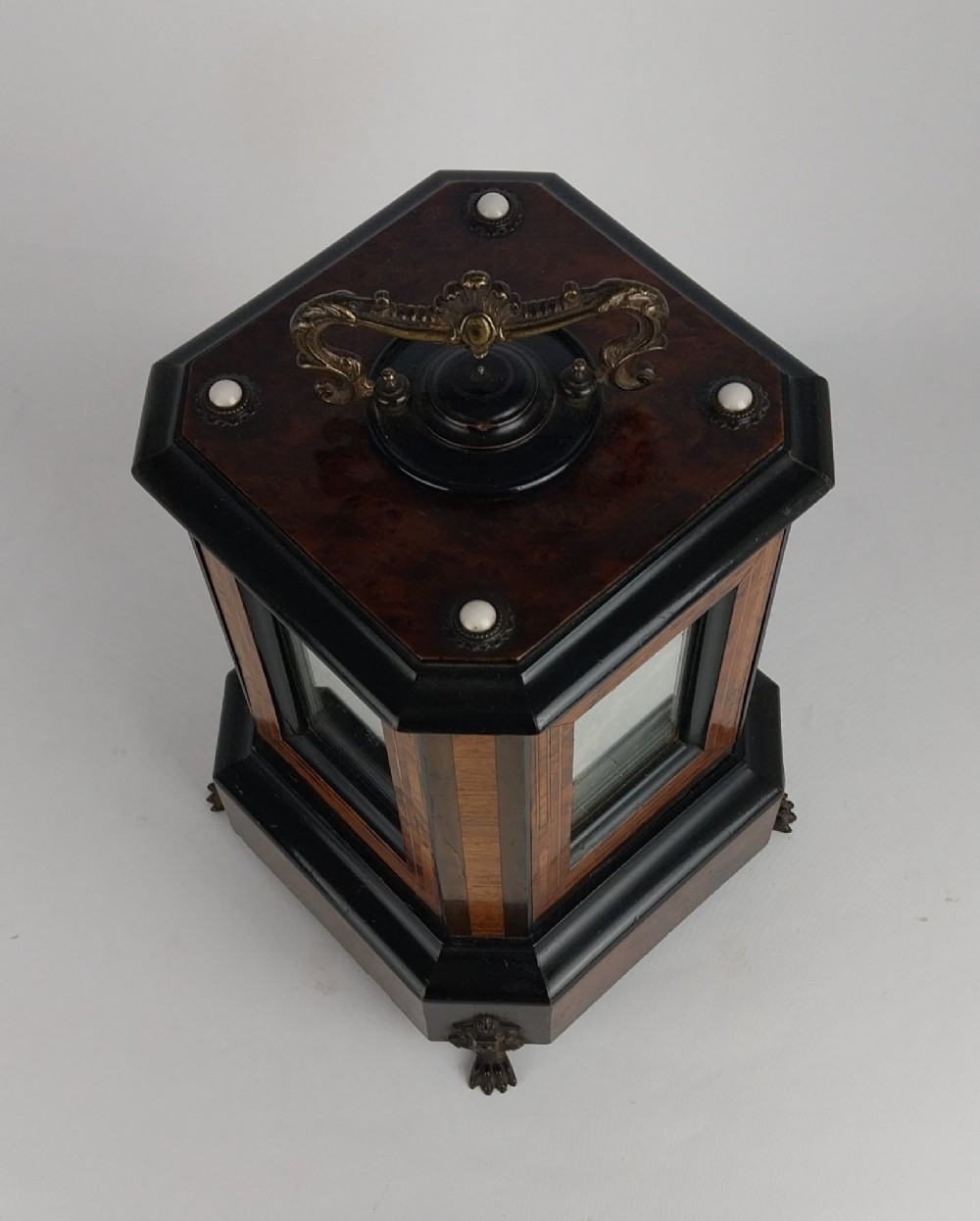 An upright Victorian hexagonal musical cigar box featuring ebonised wood, burr walnut panels, glass inserts, an ornate brass top handle, and brass claw feet. View VII