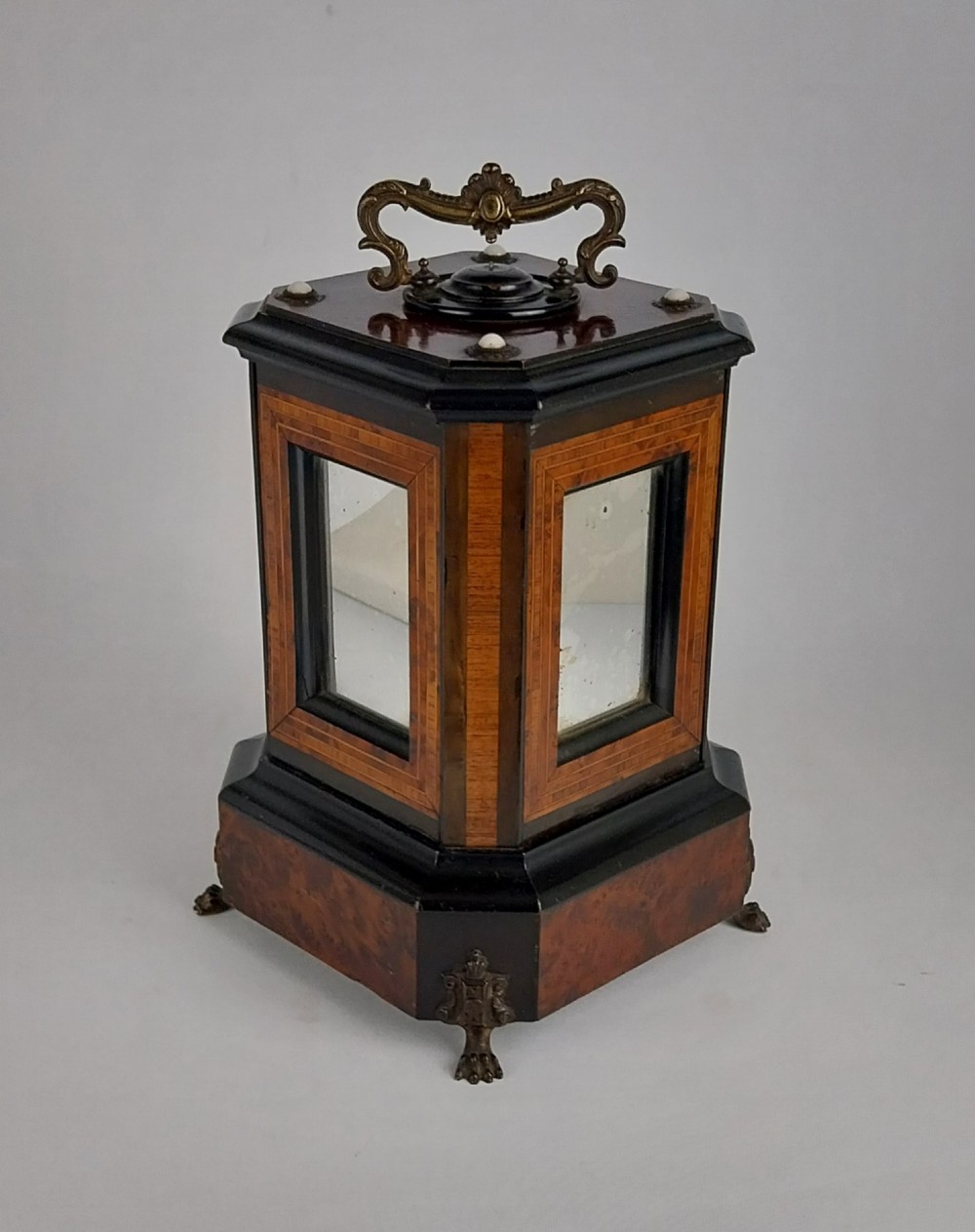 An upright Victorian hexagonal musical cigar box featuring ebonised wood, burr walnut panels, glass inserts, an ornate brass top handle, and brass claw feet. View XV