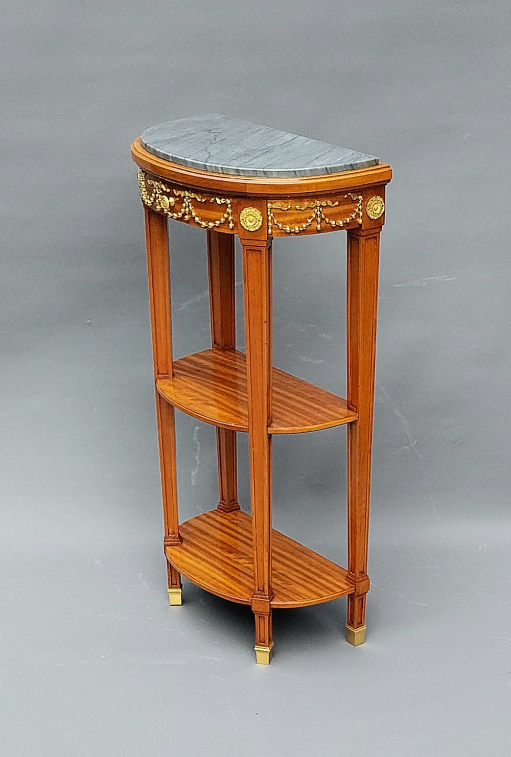 A pair of semi-circular French satinwood etagere tables featuring grey marble tops, gilt bronze garland mounts, and two lower tiers. View IX