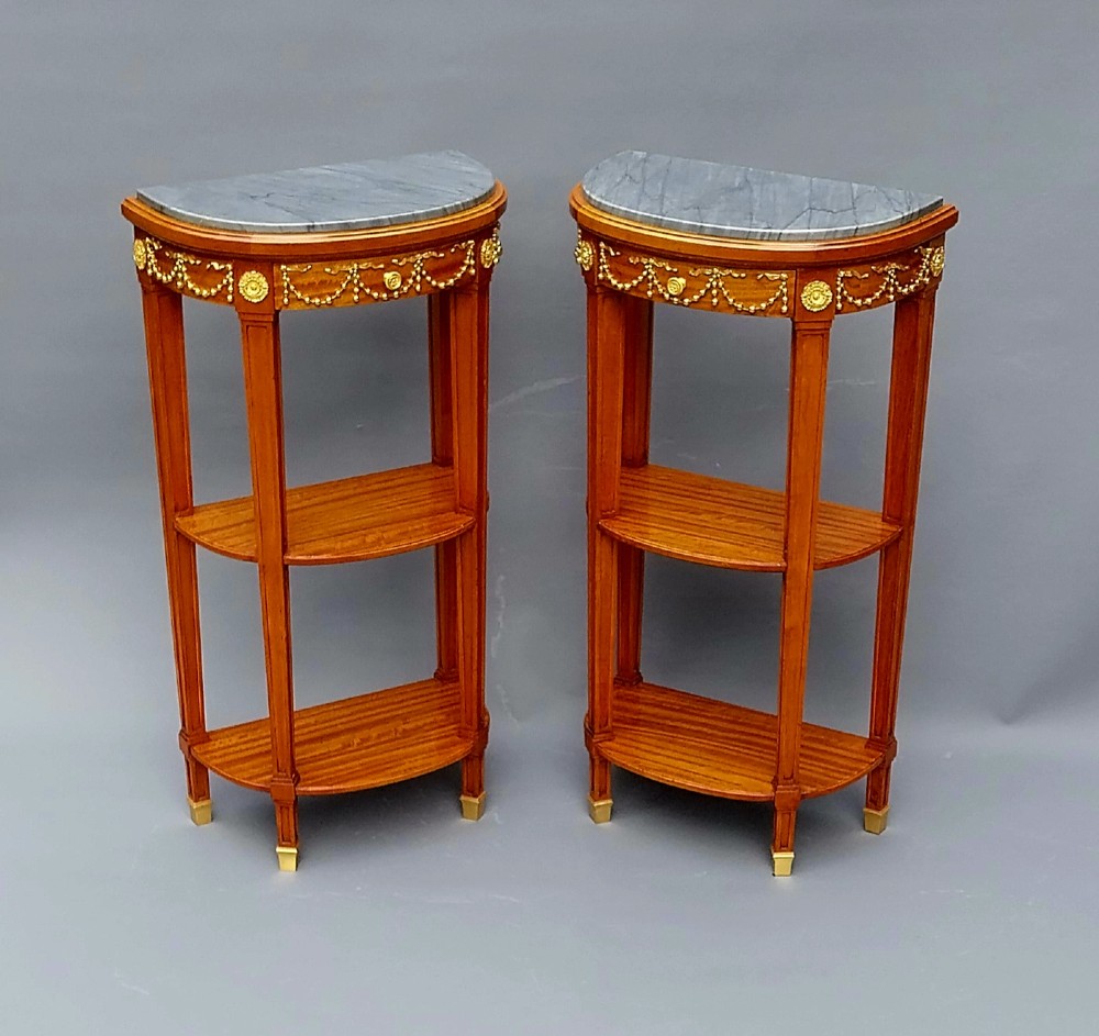 A pair of French Louis XVI style demilune side tables featuring grey marble tops, open two-tiered wooden shelving, and ornate gilt bronze floral garland mounts.