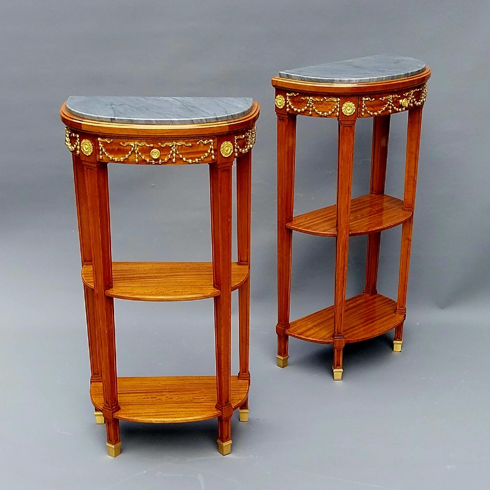 A pair of French Louis XVI style demilune side tables featuring grey marble tops, open two-tiered wooden shelving, and ornate gilt bronze floral garland mounts. View IV