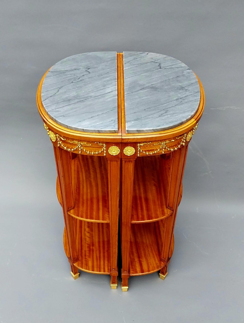 A pair of French Louis XVI style demilune side tables featuring grey marble tops, open two-tiered wooden shelving, and ornate gilt bronze floral garland mounts. View V
