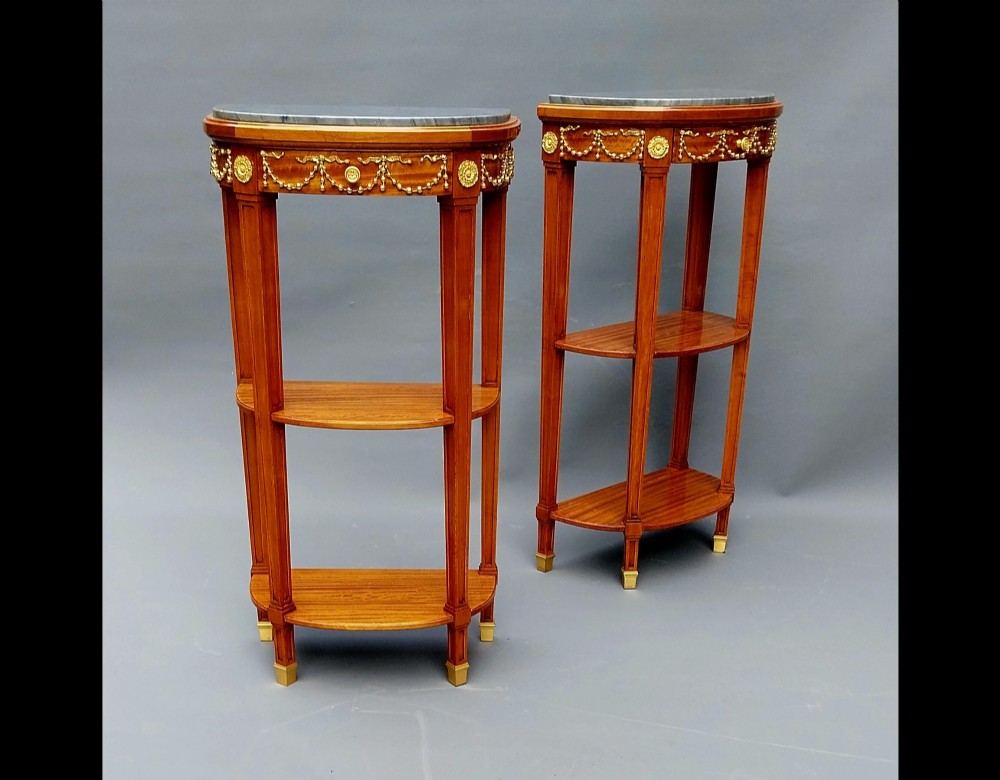A pair of French Louis XVI style demilune side tables featuring grey marble tops, open two-tiered wooden shelving, and ornate gilt bronze floral garland mounts. View VI