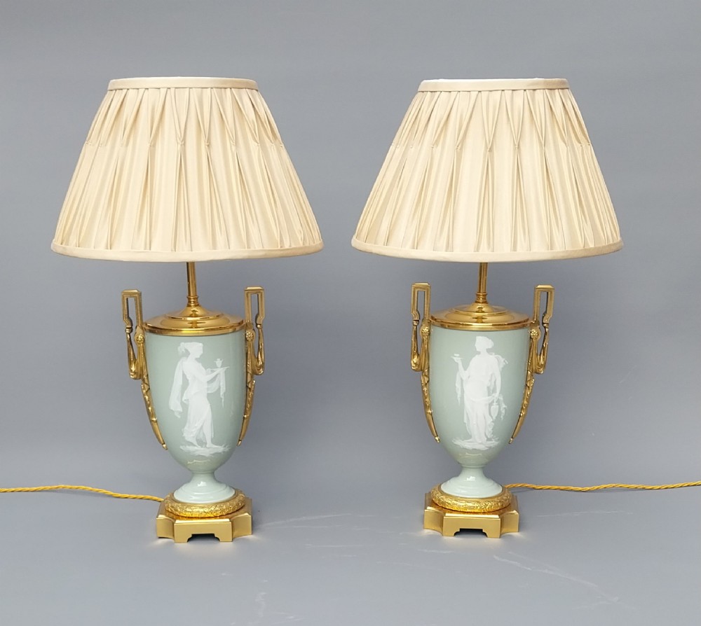 A pair of 19th-century French celadon green porcelain urn table lamps featuring white pate-sur-pate classical relief figures, gilt bronze handles and bases, and cream pleated lampshades.