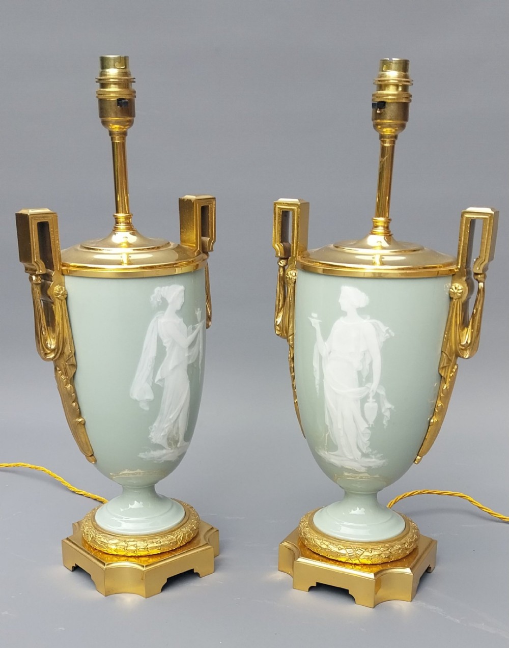 A pair of 19th-century French celadon green porcelain urn table lamps featuring white pate-sur-pate classical relief figures, gilt bronze handles and bases, and cream pleated lampshades. View V