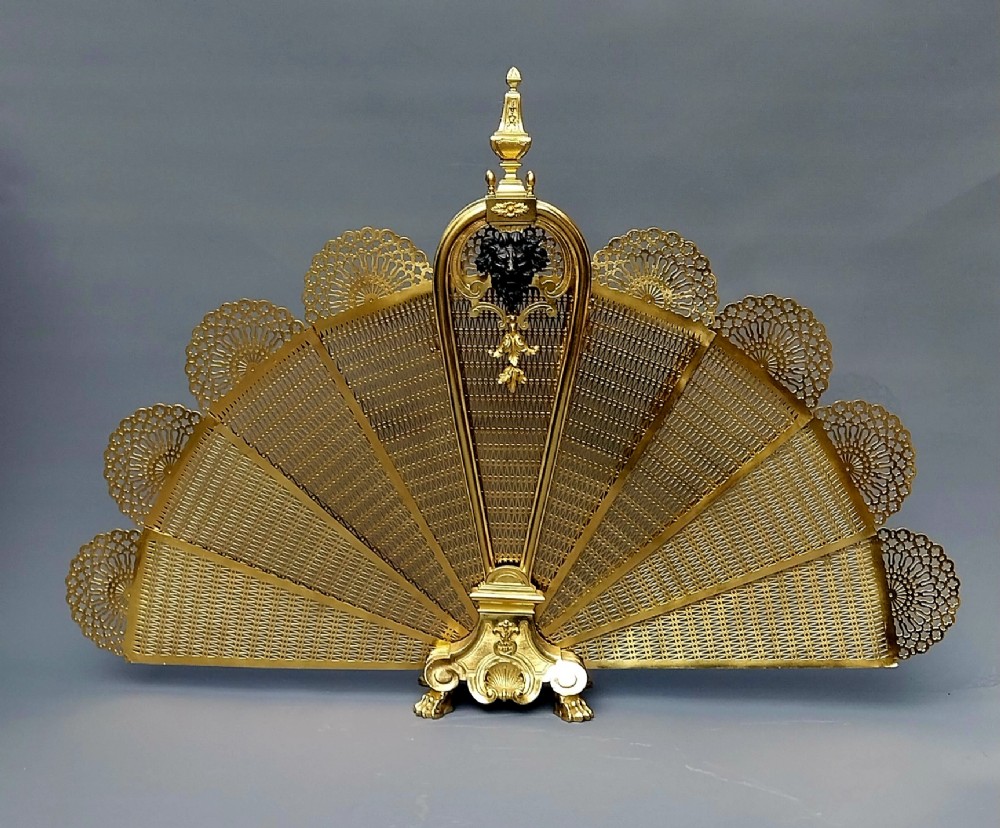 A French gilt bronze fan-shaped fire screen featuring folding filigree mesh panels, an ornate central standard with a dark mask, and a cast shell pedestal base.