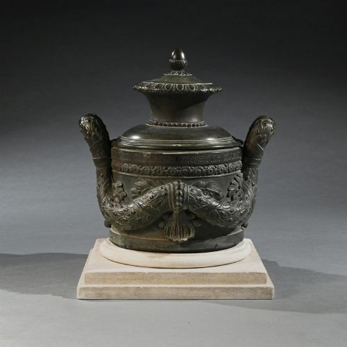 fig 1. A dark patinated 18th-century Italian bronze vessel featuring relief garlands, handles, and Latin inscriptions, resting on a beige stepped stone plinth.