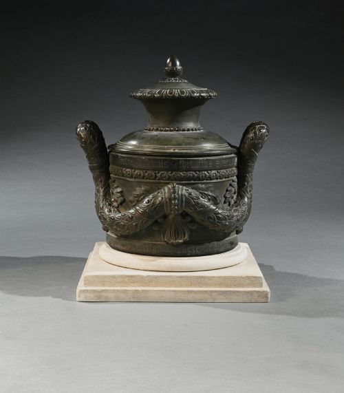 fig 2. A dark patinated 18th-century Italian bronze vessel featuring relief garlands, handles, and Latin inscriptions, resting on a beige stepped stone plinth.