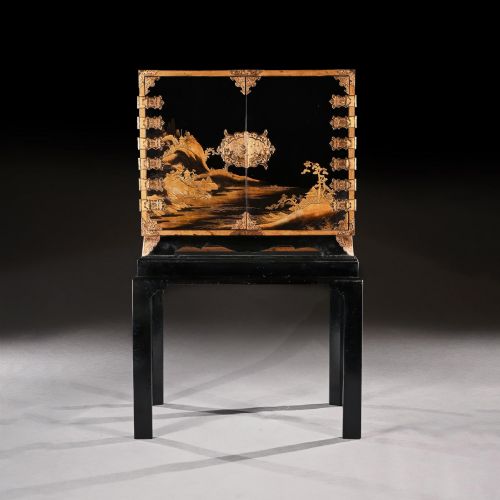 fig 3. A black lacquered Japanese cabinet decorated with gold maki-e landscape scenes, featuring ornate brass mounts and elevated on a simple black stand.