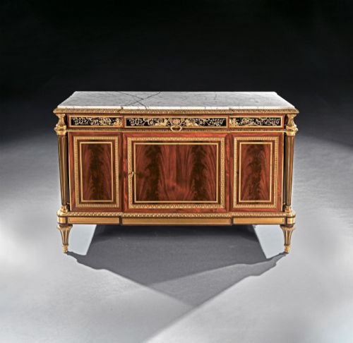 fig 1. A 19th-century French Louis XVI style flame mahogany commode featuring ornate gilt-bronze mounts, three paneled cupboard doors, fluted legs, and a white marble top.