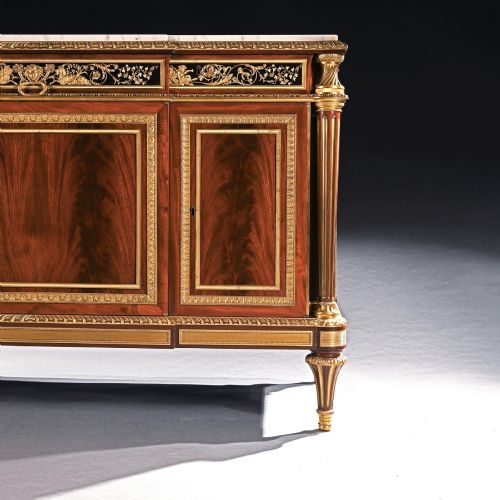fig 3. A 19th-century French Louis XVI style flame mahogany commode featuring ornate gilt-bronze mounts, three paneled cupboard doors, fluted legs, and a white marble top.
