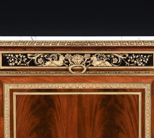 fig 4. A 19th-century French Louis XVI style flame mahogany commode featuring ornate gilt-bronze mounts, three paneled cupboard doors, fluted legs, and a white marble top.