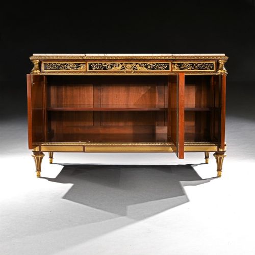 fig 5. A 19th-century French Louis XVI style flame mahogany commode featuring ornate gilt-bronze mounts, three paneled cupboard doors, fluted legs, and a white marble top.