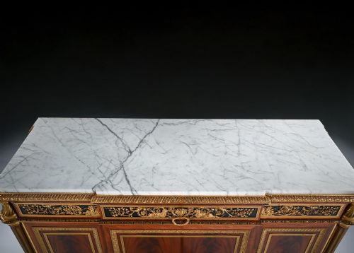 fig 6. A 19th-century French Louis XVI style flame mahogany commode featuring ornate gilt-bronze mounts, three paneled cupboard doors, fluted legs, and a white marble top.