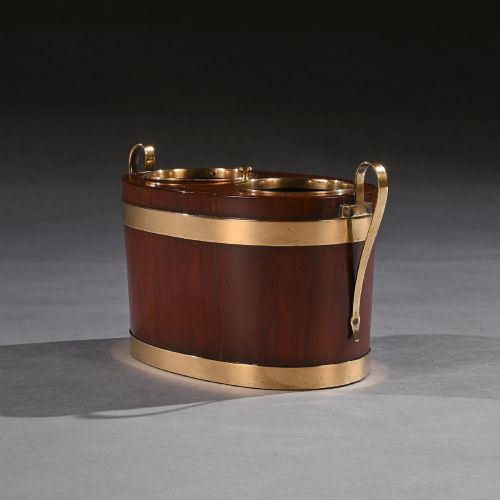 fig 1. An oval George III mahogany table wine cooler featuring polished brass bands, two top bottle openings, a central finial, and elevated brass side handles.