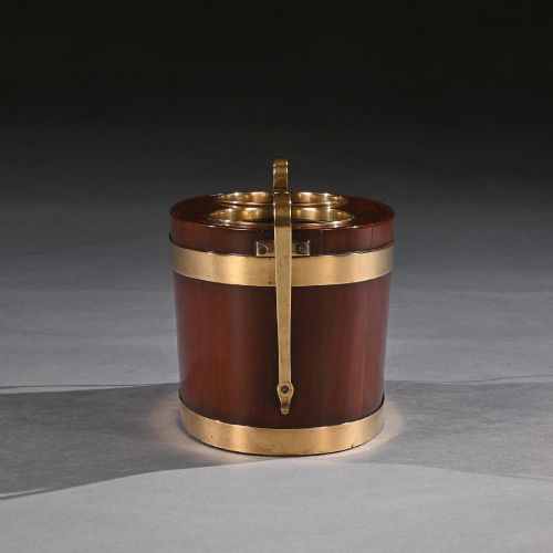 fig 3. An oval George III mahogany table wine cooler featuring polished brass bands, two top bottle openings, a central finial, and elevated brass side handles.