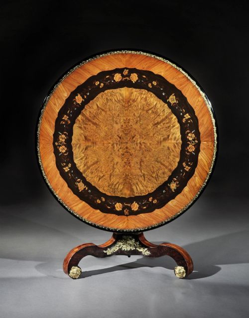 fig 1. A 19th-century circular marquetry centre table featuring floral wood inlays, a tilt-top mechanism, and a dark carved tripod base with gilt-bronze mounts.