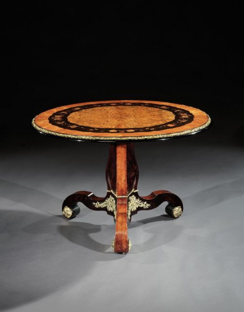 fig 2. A 19th-century circular marquetry centre table featuring floral wood inlays, a tilt-top mechanism, and a dark carved tripod base with gilt-bronze mounts.