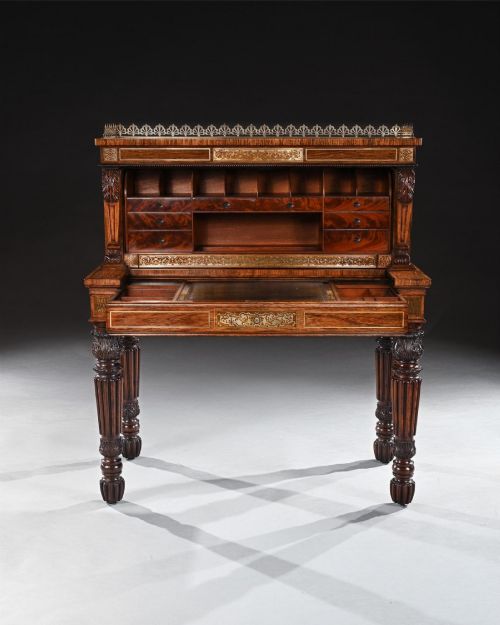 fig 1. A Regency rosewood writing table featuring intricate brass scrollwork inlay, an anthemion top gallery, a central frieze drawer, and carved reeded legs.