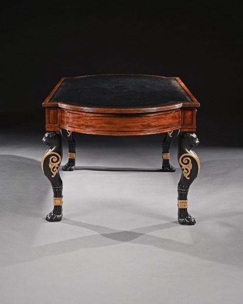 fig 2. A Regency style mahogany writing table featuring a dark leather inset top, frieze drawers, and four ebonised monopodia legs ornamented with carved lion heads and gilt accents.