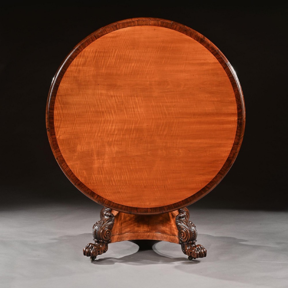 A nineteenth-century circular satinwood centre table featuring rosewood crossbanding, a central pedestal support, and a carved triform base with paw feet on castors. View II