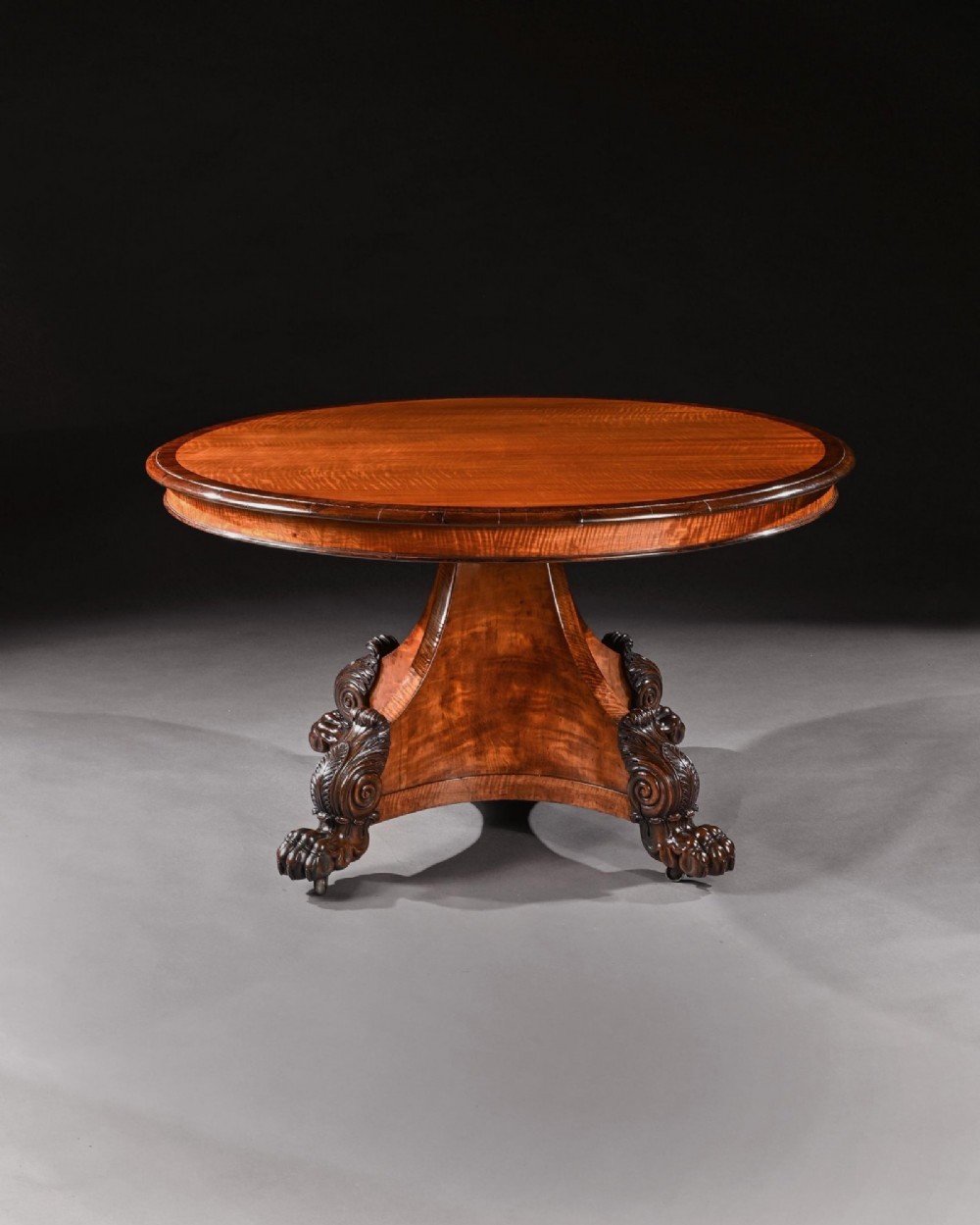A nineteenth-century circular satinwood centre table featuring rosewood crossbanding, a central pedestal support, and a carved triform base with paw feet on castors. View III