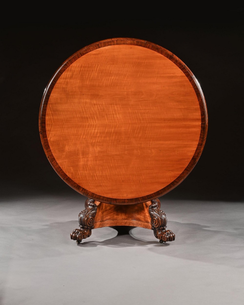 A nineteenth-century circular satinwood centre table featuring rosewood crossbanding, a central pedestal support, and a carved triform base with paw feet on castors. View IV