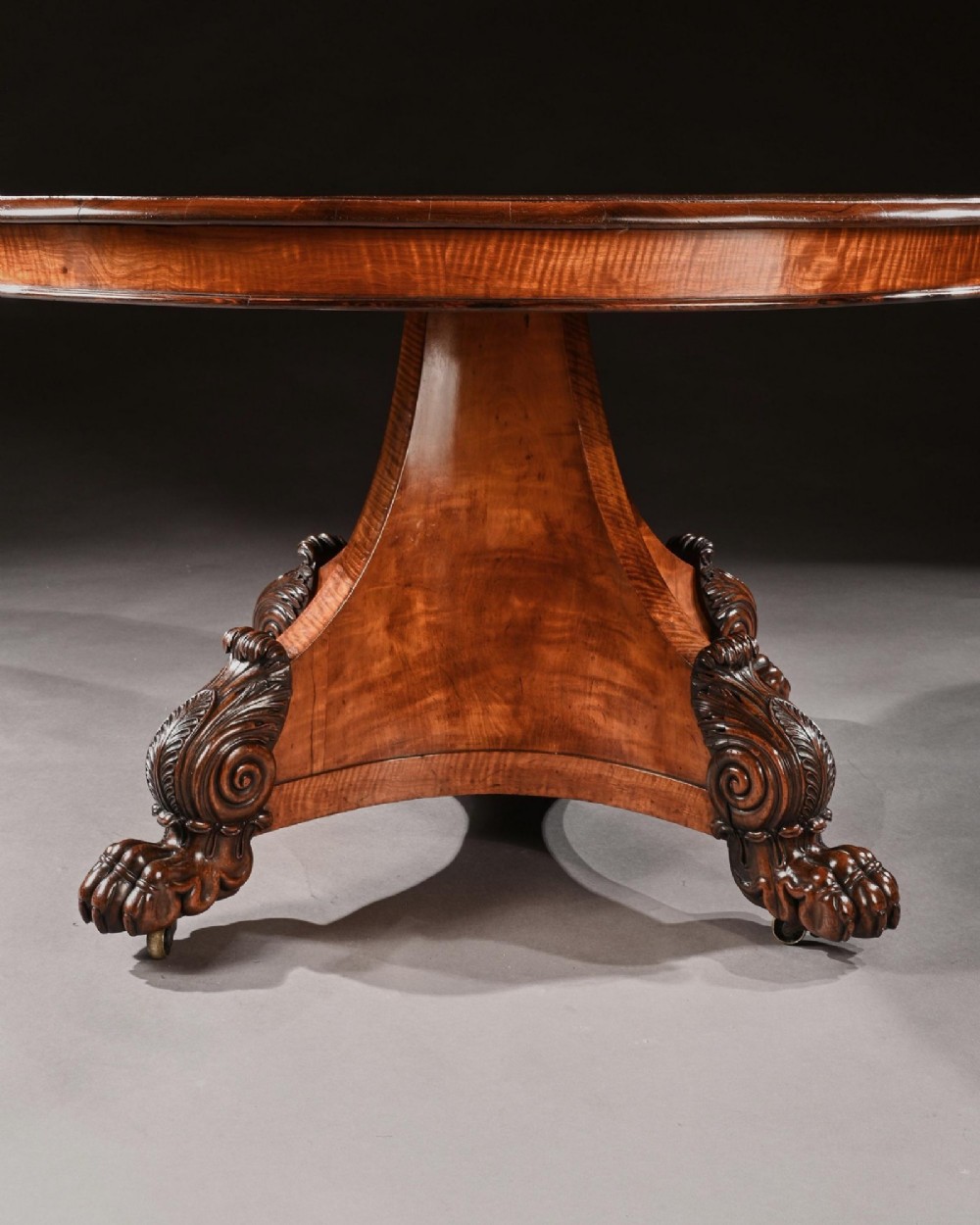 A nineteenth-century circular satinwood centre table featuring rosewood crossbanding, a central pedestal support, and a carved triform base with paw feet on castors. View V