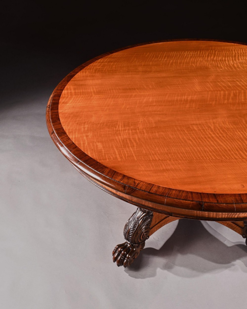 A nineteenth-century circular satinwood centre table featuring rosewood crossbanding, a central pedestal support, and a carved triform base with paw feet on castors. View VII