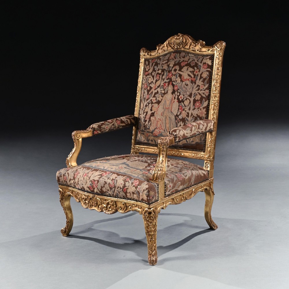 An 18th-century French Regence giltwood armchair featuring ornate shell carving, cabriole legs, and tapestry upholstery depicting figures and floral motifs.