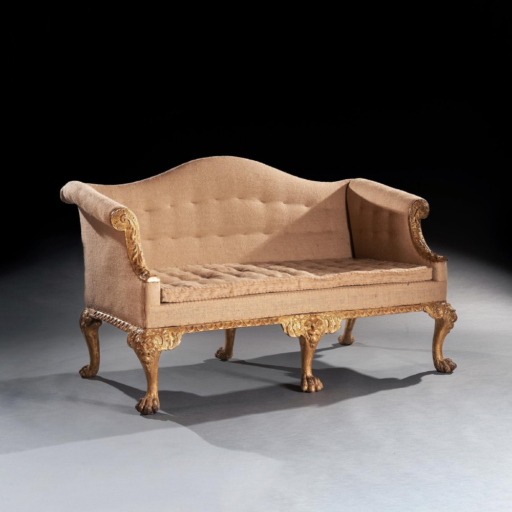 A George II carved giltwood sofa upholstered in neutral beige fabric, supported on six cabriole legs featuring carved lion mask knees and hairy paw feet.
