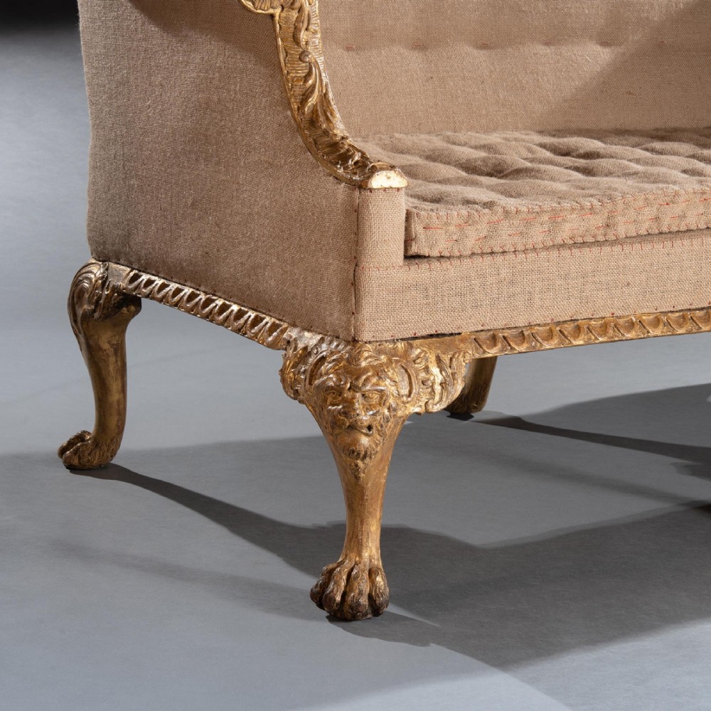 A George II carved giltwood sofa upholstered in neutral beige fabric, supported on six cabriole legs featuring carved lion mask knees and hairy paw feet. View IV