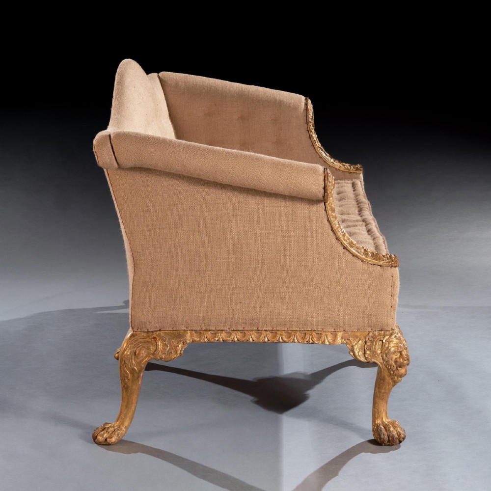 A George II carved giltwood sofa upholstered in neutral beige fabric, supported on six cabriole legs featuring carved lion mask knees and hairy paw feet. View IX