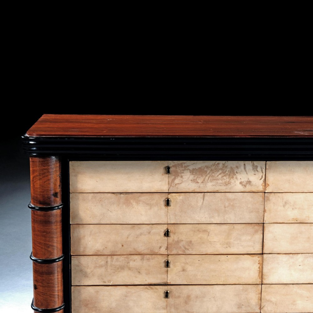 An Art Deco palisander commode featuring twelve parchment-covered drawer fronts, ebonised side trim, curved timber flanks, and a stepped plinth base. View IV