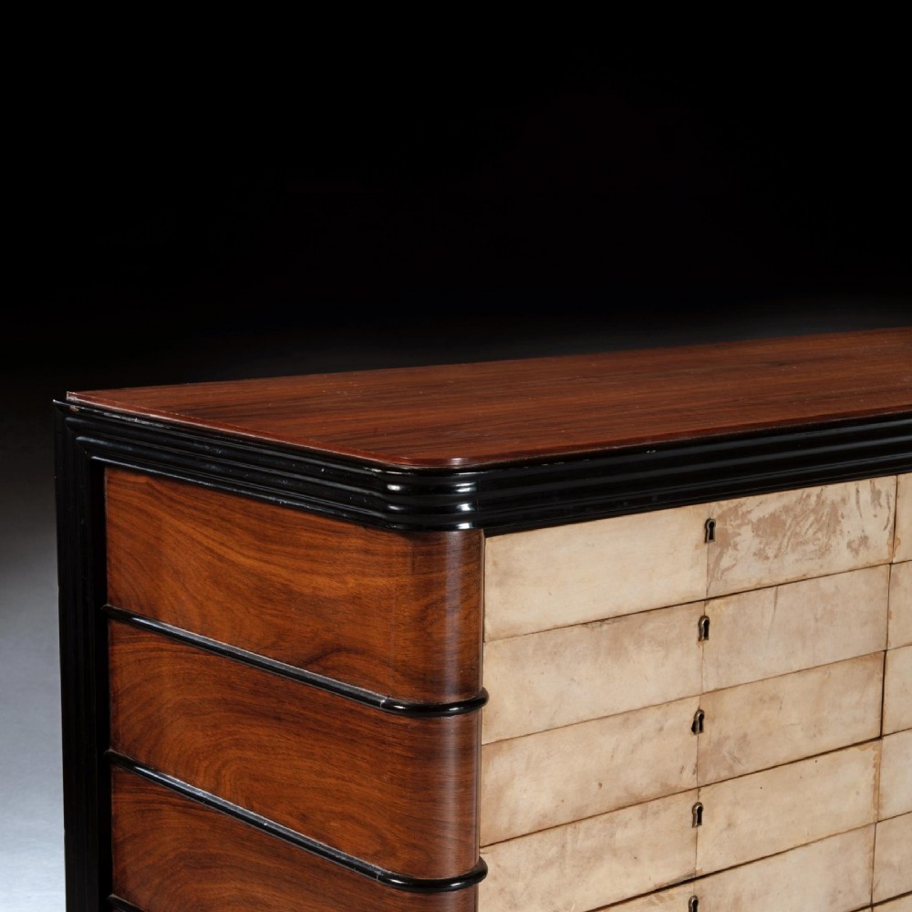 An Art Deco palisander commode featuring twelve parchment-covered drawer fronts, ebonised side trim, curved timber flanks, and a stepped plinth base. View V
