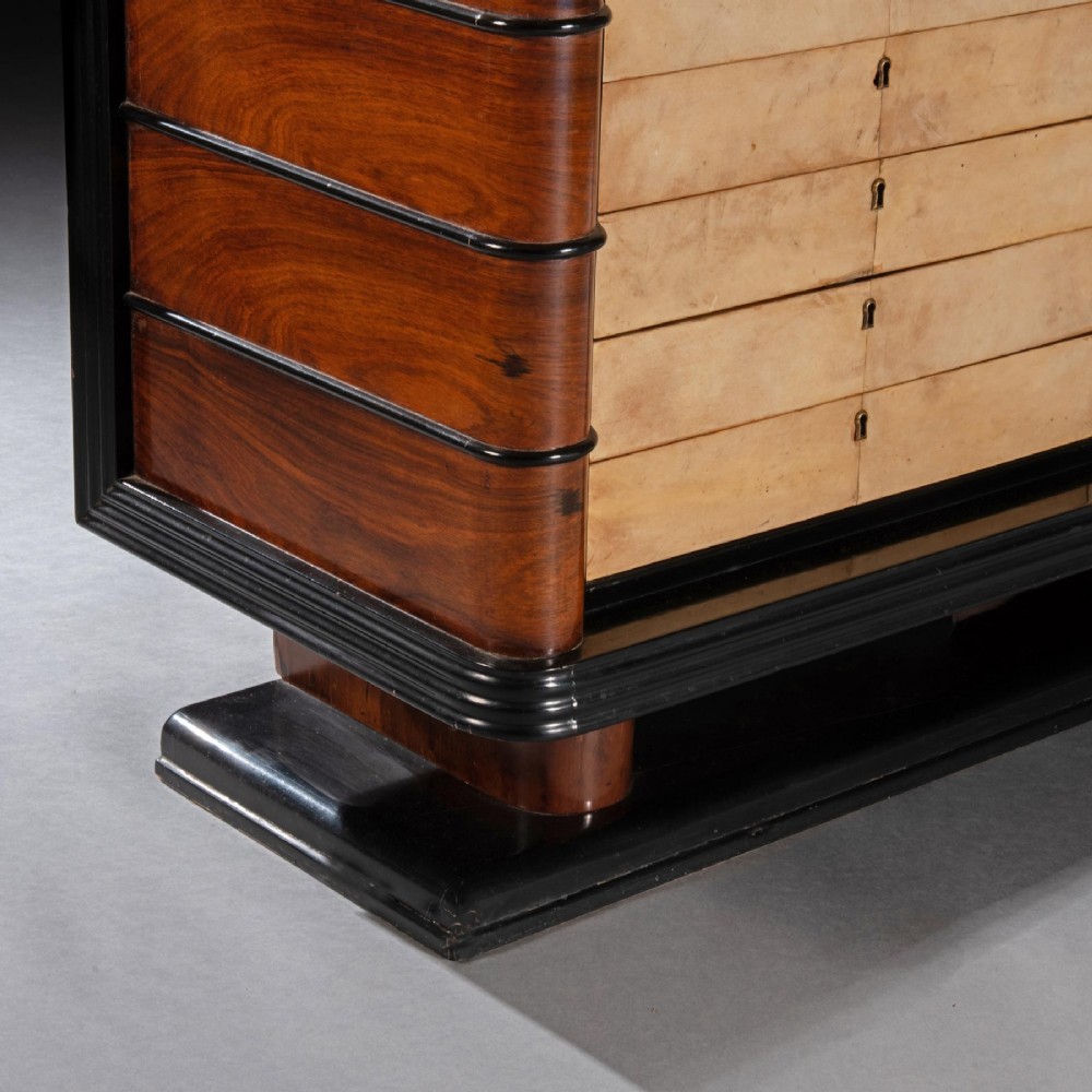 An Art Deco palisander commode featuring twelve parchment-covered drawer fronts, ebonised side trim, curved timber flanks, and a stepped plinth base. View VI