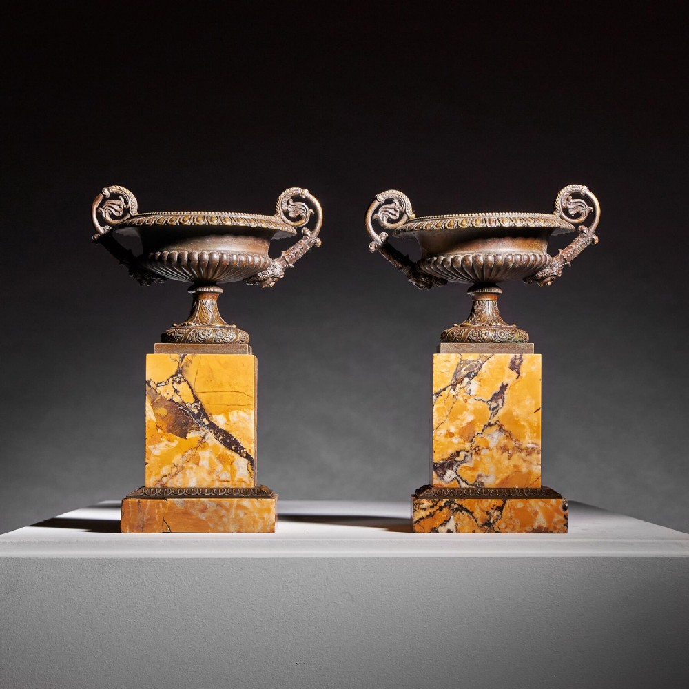 A pair of early nineteenth-century French patinated bronze tazzas with ornate scroll handles mounted on square veined yellow marble plinths.